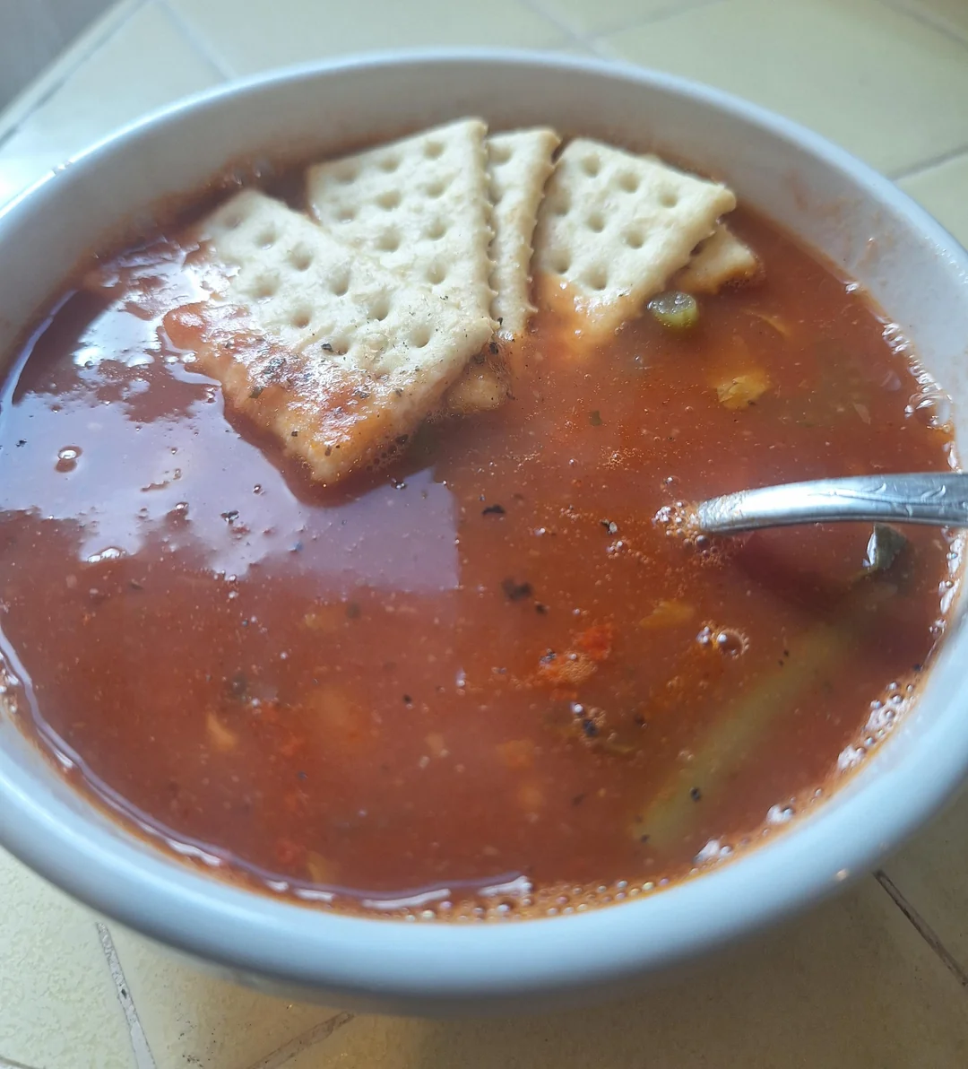 A bowl of tomato-based soup with vegetables, topped with several square saltine crackers. A metal spoon is resting in the soup, and the bowl is placed on a light-colored tiled surface.