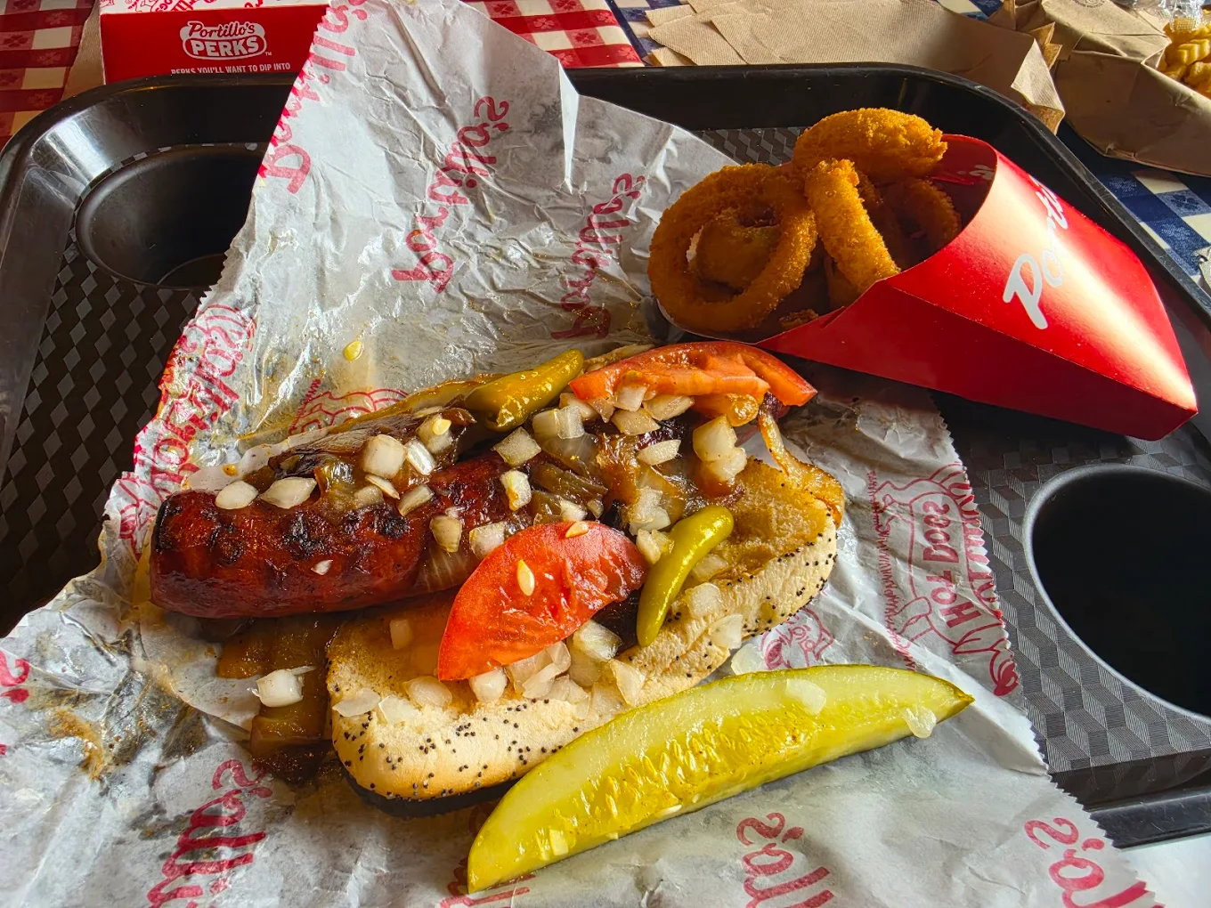 A grilled sausage on a poppy seed bun topped with onions, peppers, and tomato slices, served with a dill pickle spear and a side of onion rings on a black tray.