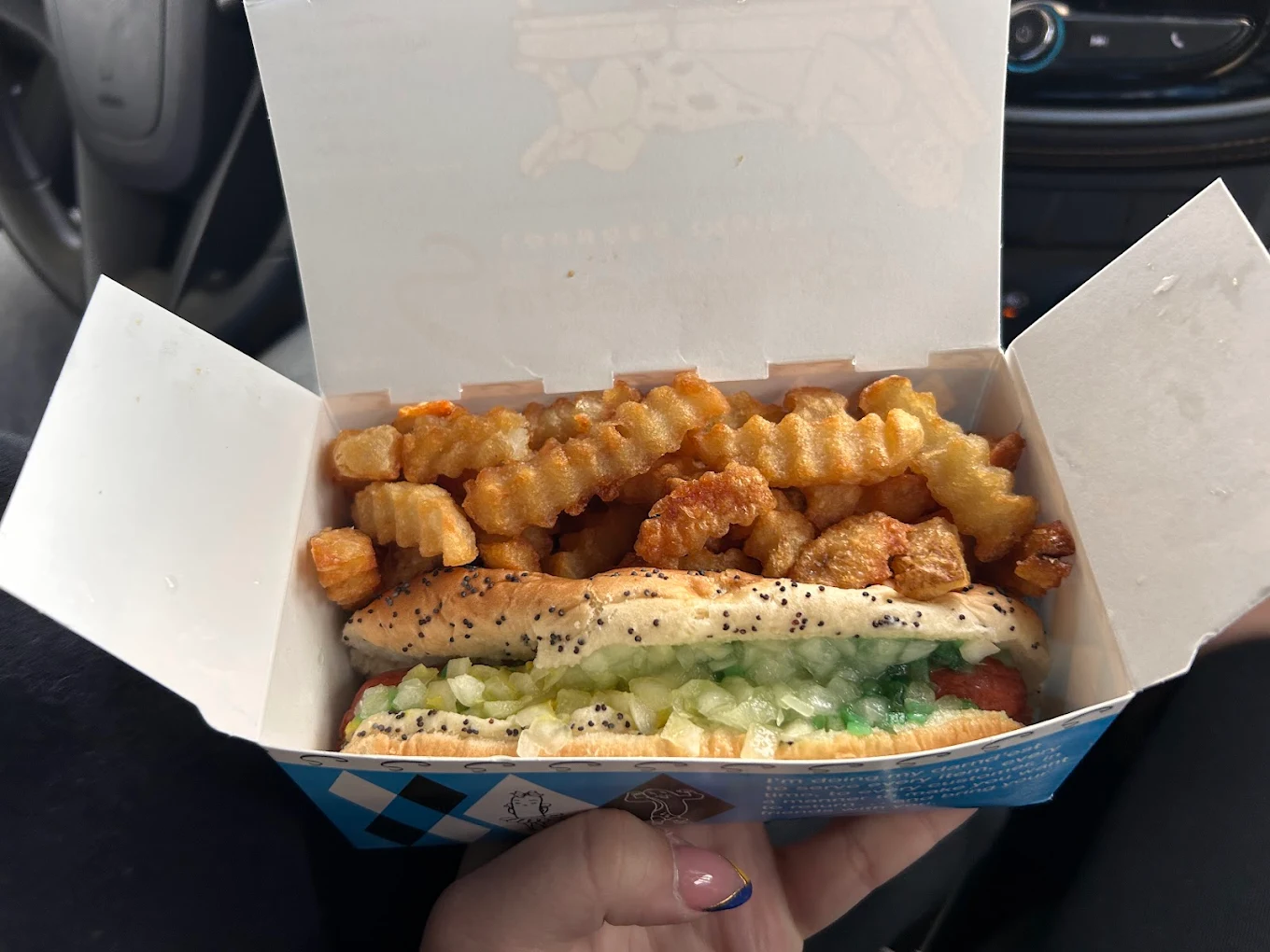 A hand holds an open box containing a poppy seed bun hot dog topped with chopped onions and relish, alongside crinkle-cut fries and fried cheese curds. The background shows part of a car interior.