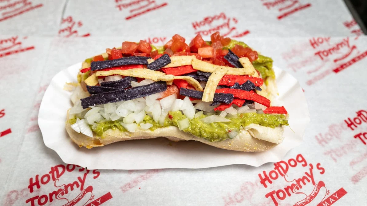 A hot dog topped with chopped onions, guacamole, diced tomatoes, and colorful tortilla strips on a paper tray, placed on paper printed with "Hot Dog Tommy's.