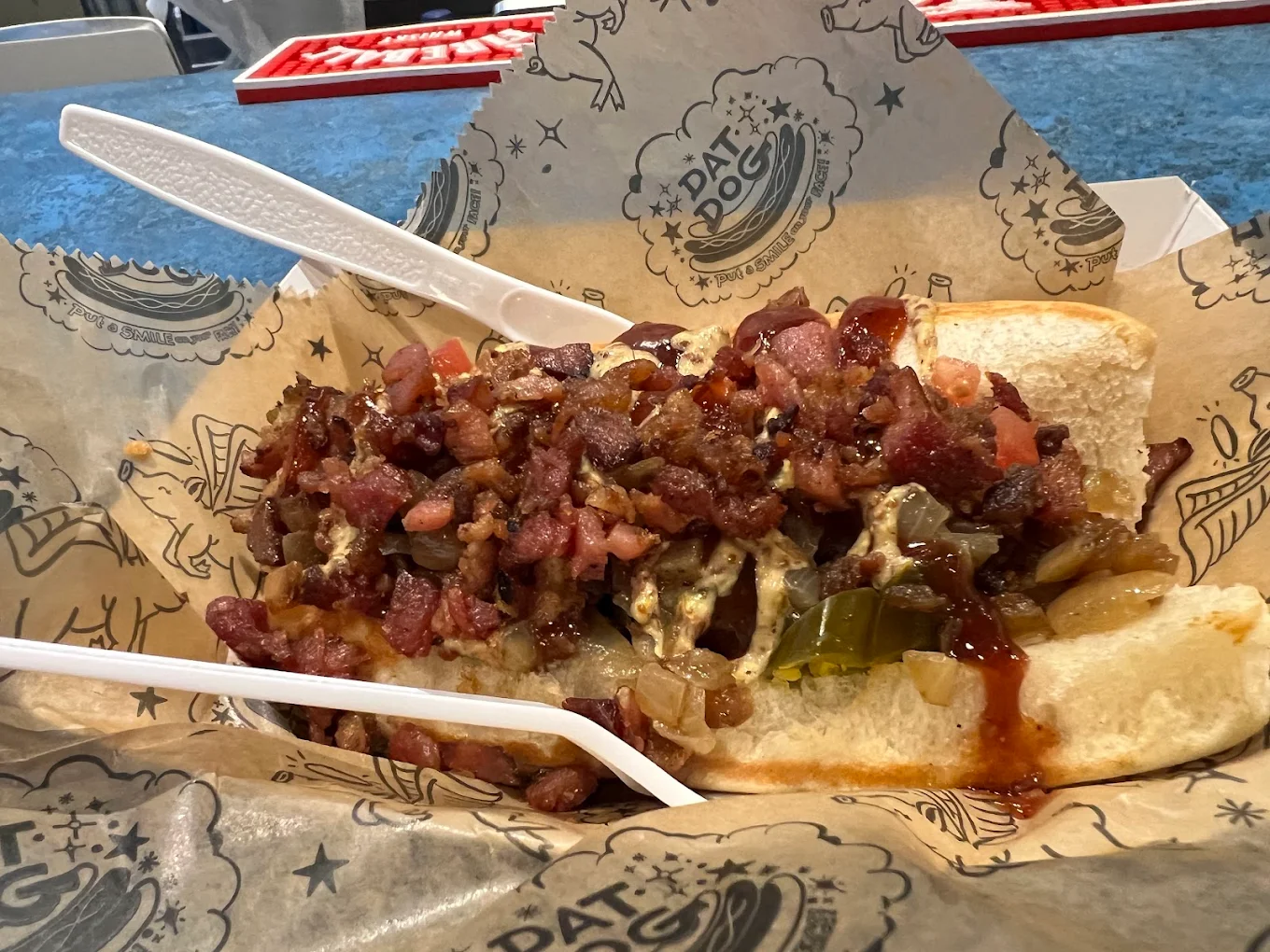 A hot dog in a bun topped with chopped bacon, grilled onions, pickles, and barbecue sauce, served on branded parchment paper with a plastic fork and knife.
