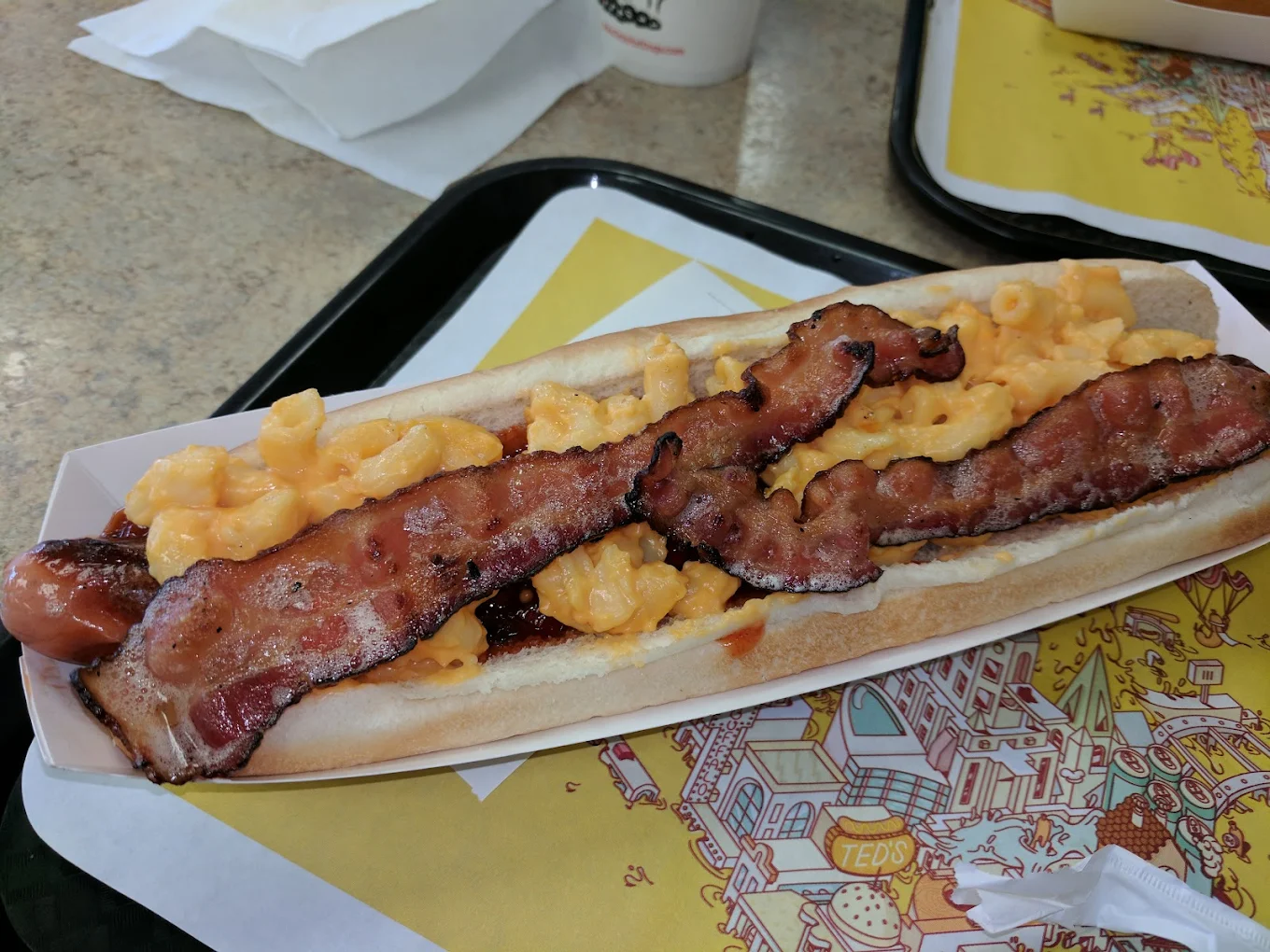 A hot dog in a tray is topped with creamy macaroni and cheese and two strips of crispy bacon, sitting on a placemat with colorful illustrations.