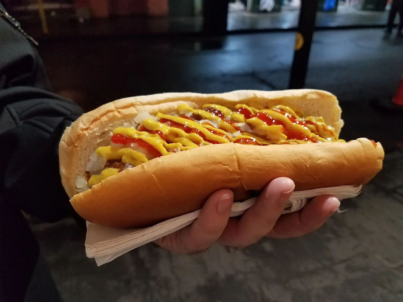 A hand holding a large hot dog topped with chopped onions, mustard, and ketchup, with the hot dog resting on a stack of napkins. The background appears to be an outdoor urban setting at night.