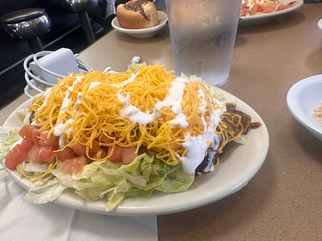 A plate of taco salad topped with shredded cheddar cheese, diced tomatoes, lettuce, sour cream, and ground beef sits on a restaurant table next to a glass of water and other dishes.