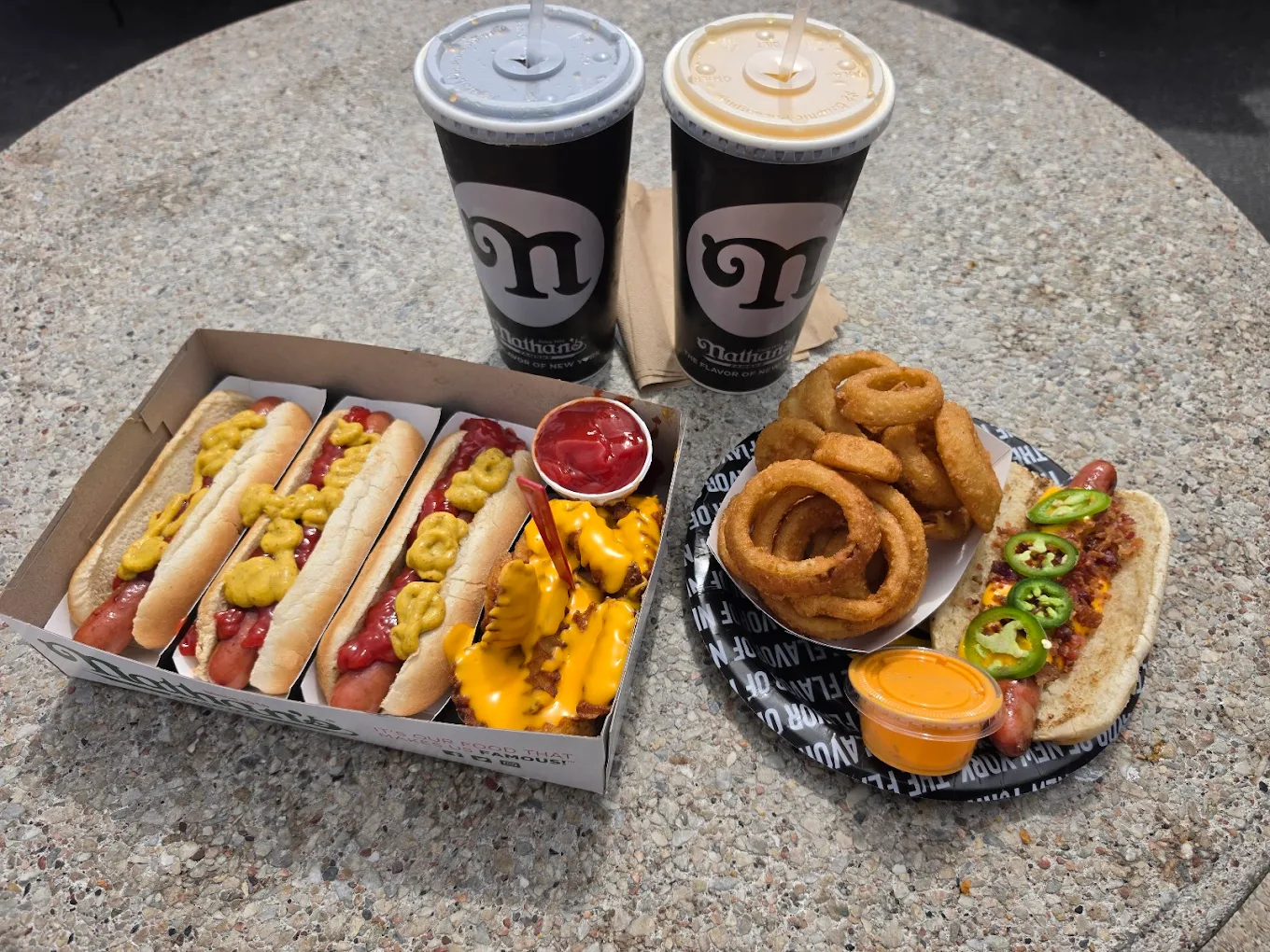 A table with three hot dogs topped with mustard in a box, a side of fries with cheese and ketchup, onion rings on a plate with a hot dog topped with jalapeños and sauce, and two large soft drinks.