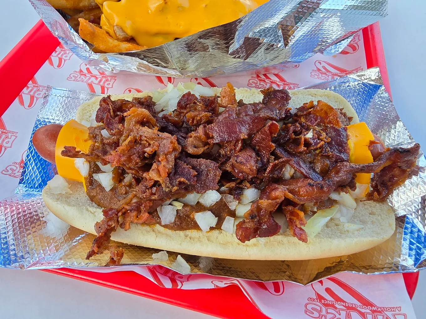 A hot dog topped with crispy bacon pieces, diced onions, and cheddar cheese on a bun, served on foil and a red tray with a side of cheese-covered fries in the background.