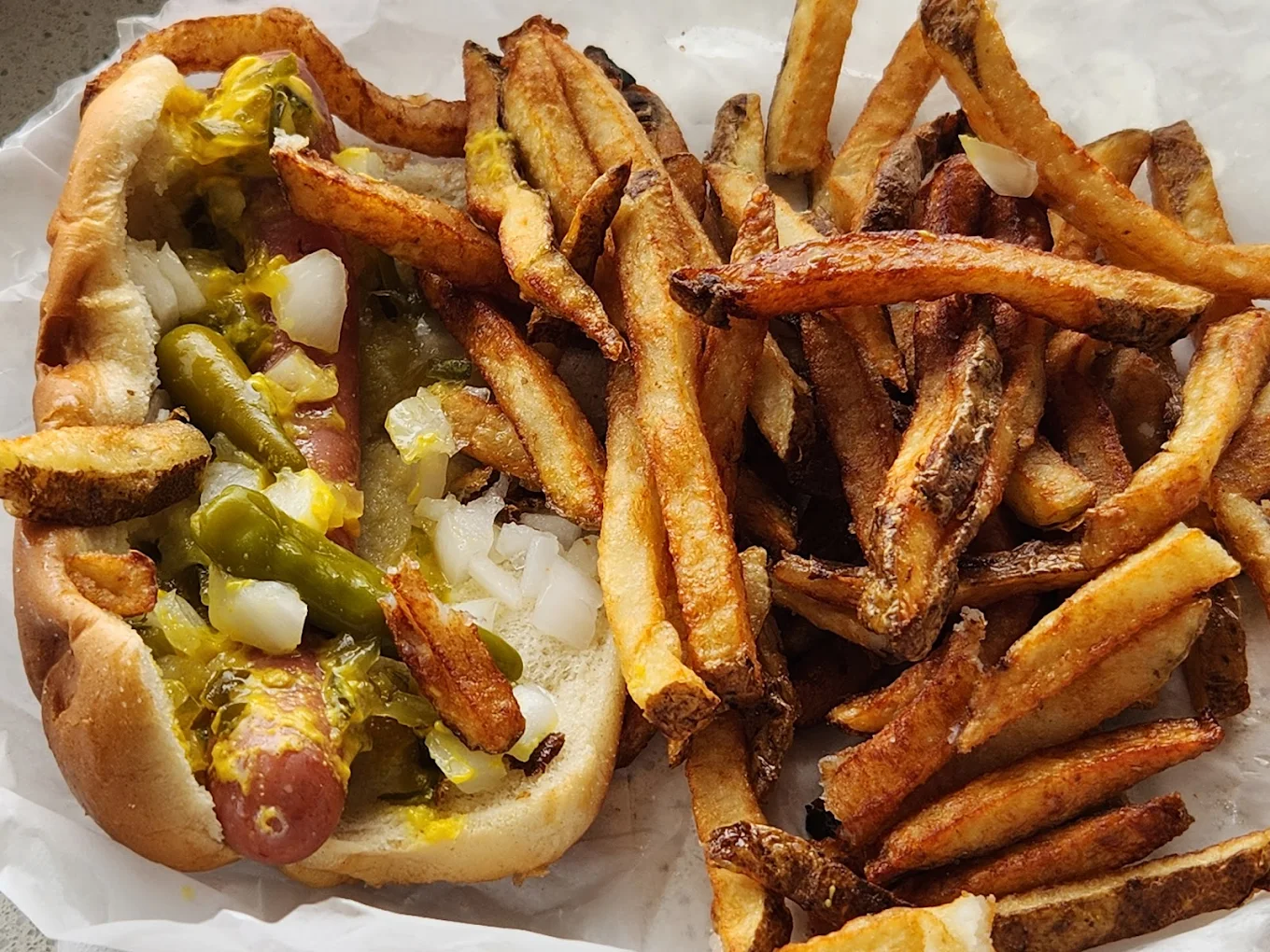 A hot dog topped with chopped onions, pickles, and mustard sits next to a generous serving of crispy golden-brown French fries on white parchment paper.