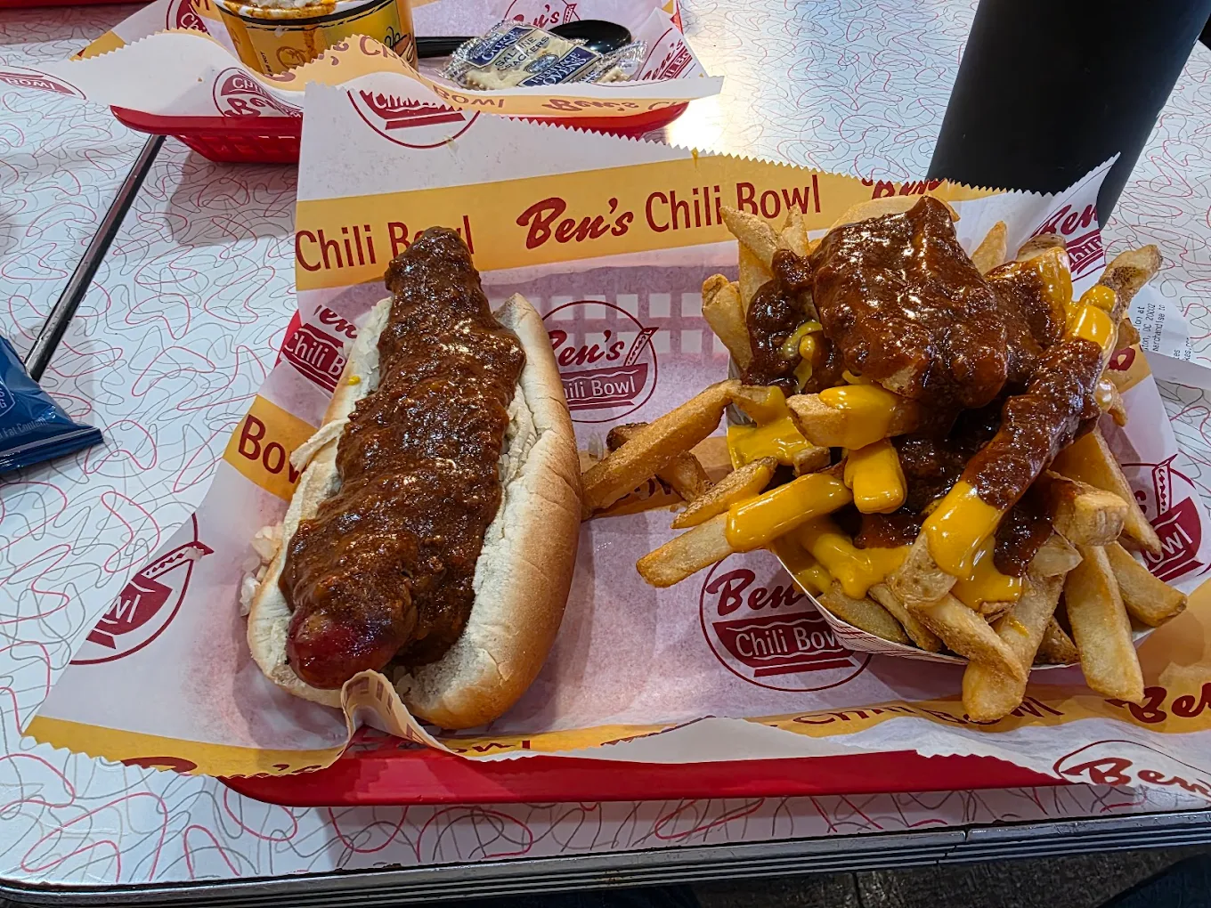 A tray with a chili dog in a bun and a serving of cheese fries topped with chili, both on Ben’s Chili Bowl branded paper. The background shows more trays of food and a patterned table surface.
