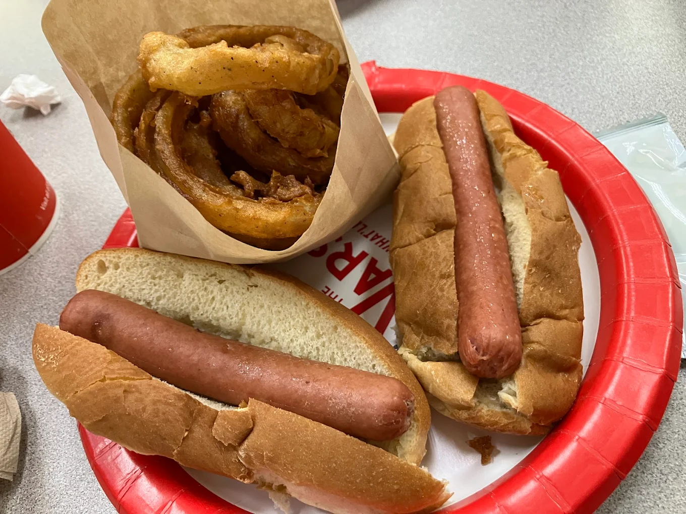Two plain hot dogs in buns and a paper cup of onion rings sit on a red and white plate on a gray tabletop. A red cup and some napkins are partially visible nearby.