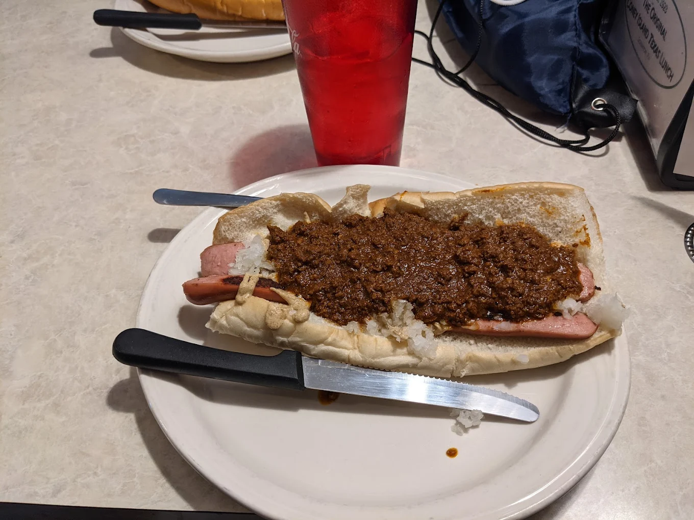 A hot dog topped with chili and chopped onions sits on a white plate with a knife and fork. A red drink cup and other items are visible in the background on a light-colored table.