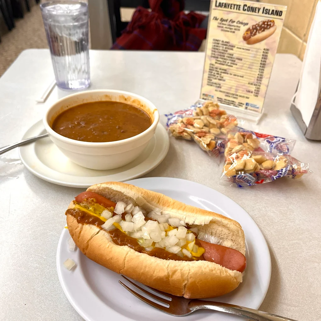A coney dog with onions, mustard, and chili sits on a plate next to a fork. A bowl of chili, packets of oyster crackers, a glass of water, and a menu are also on the table.