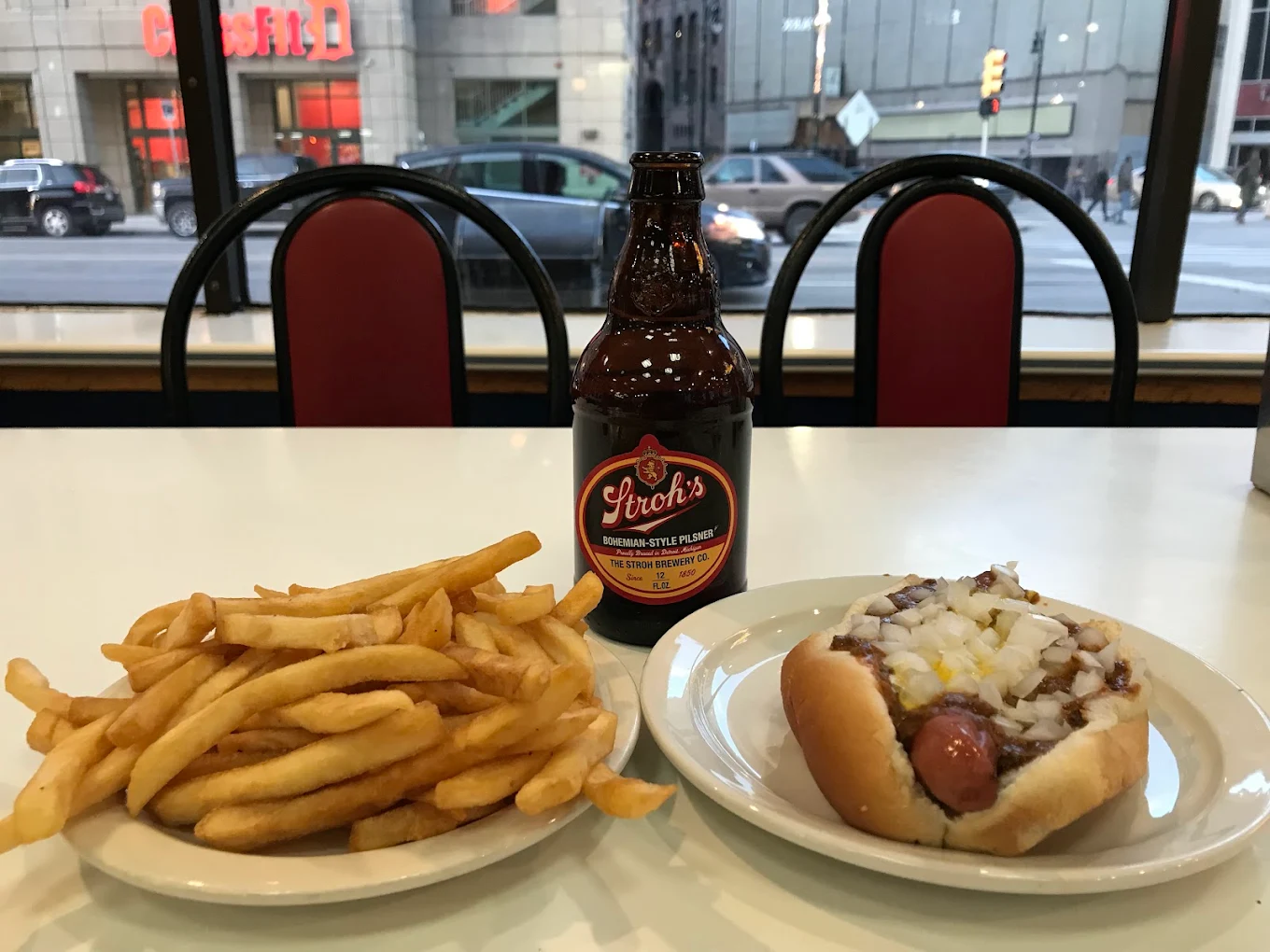 A plate of French fries, a bottle of Stroh’s beer, and a Coney Island hot dog topped with onions and chili sit on a table in a diner with large windows overlooking a city street.