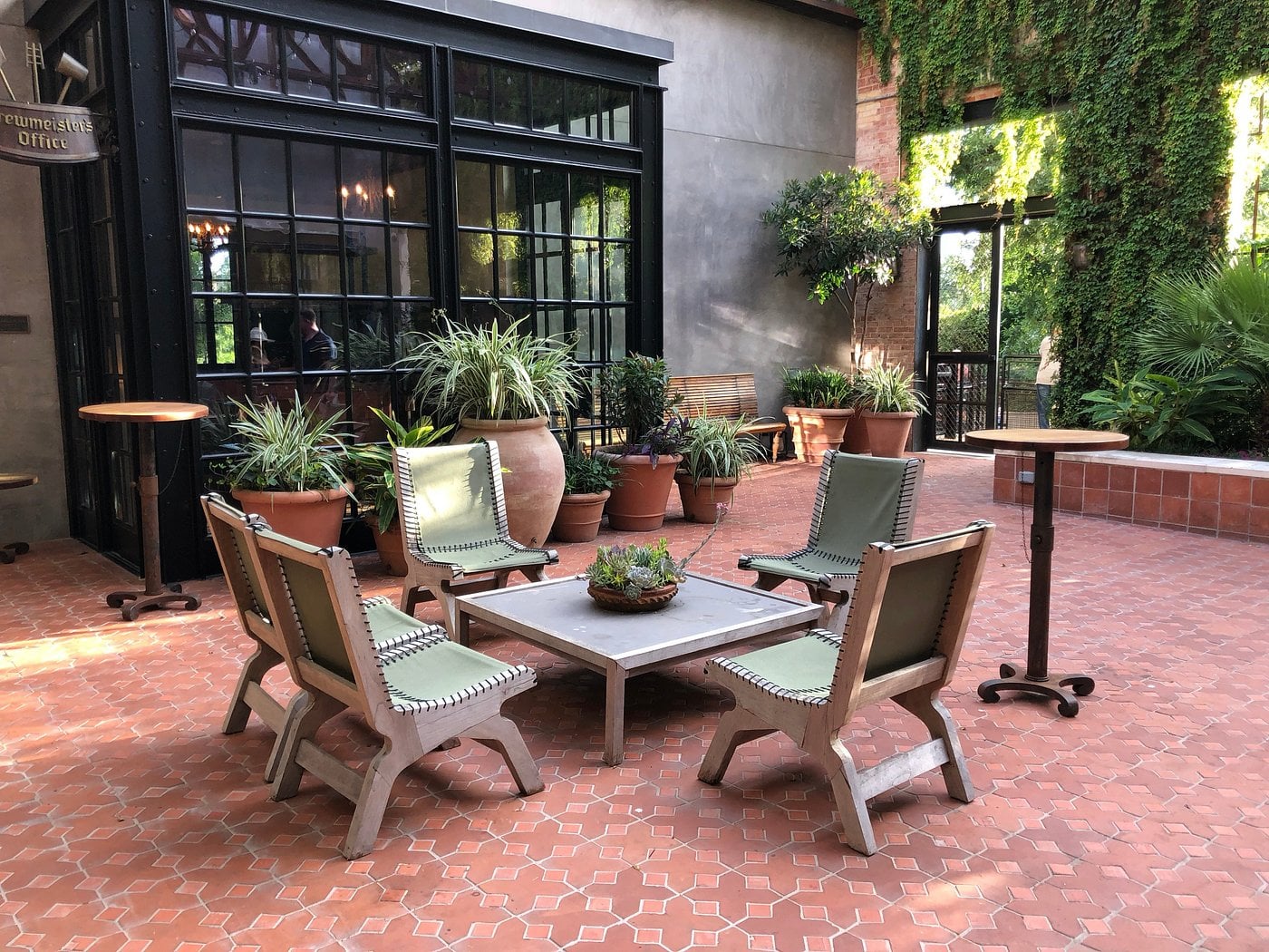 A patio with four modern chairs around a square coffee table, surrounded by potted plants and greenery on red tile flooring near a large glass wall—perfect for relaxing at Small-Town Luxury Hotels.