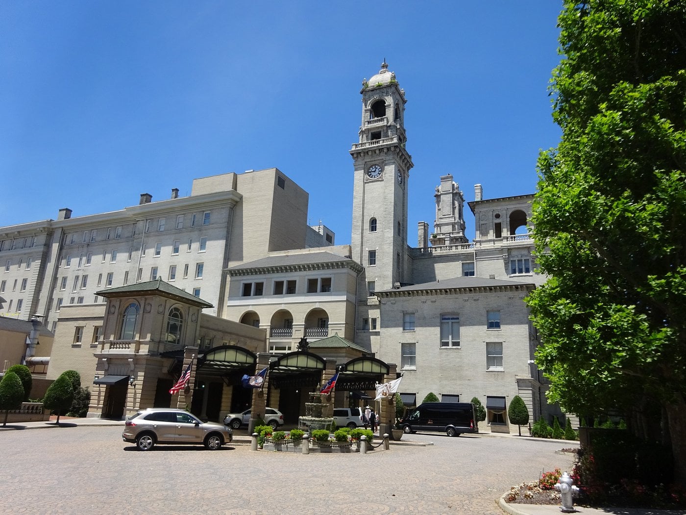 A historic, grand hotel with a tall clock tower and arched entrances embodies the charm of Small-Town Luxury Hotels. Multiple American flags fly above the main entrance, with cars parked in a circular driveway edged by manicured greenery.