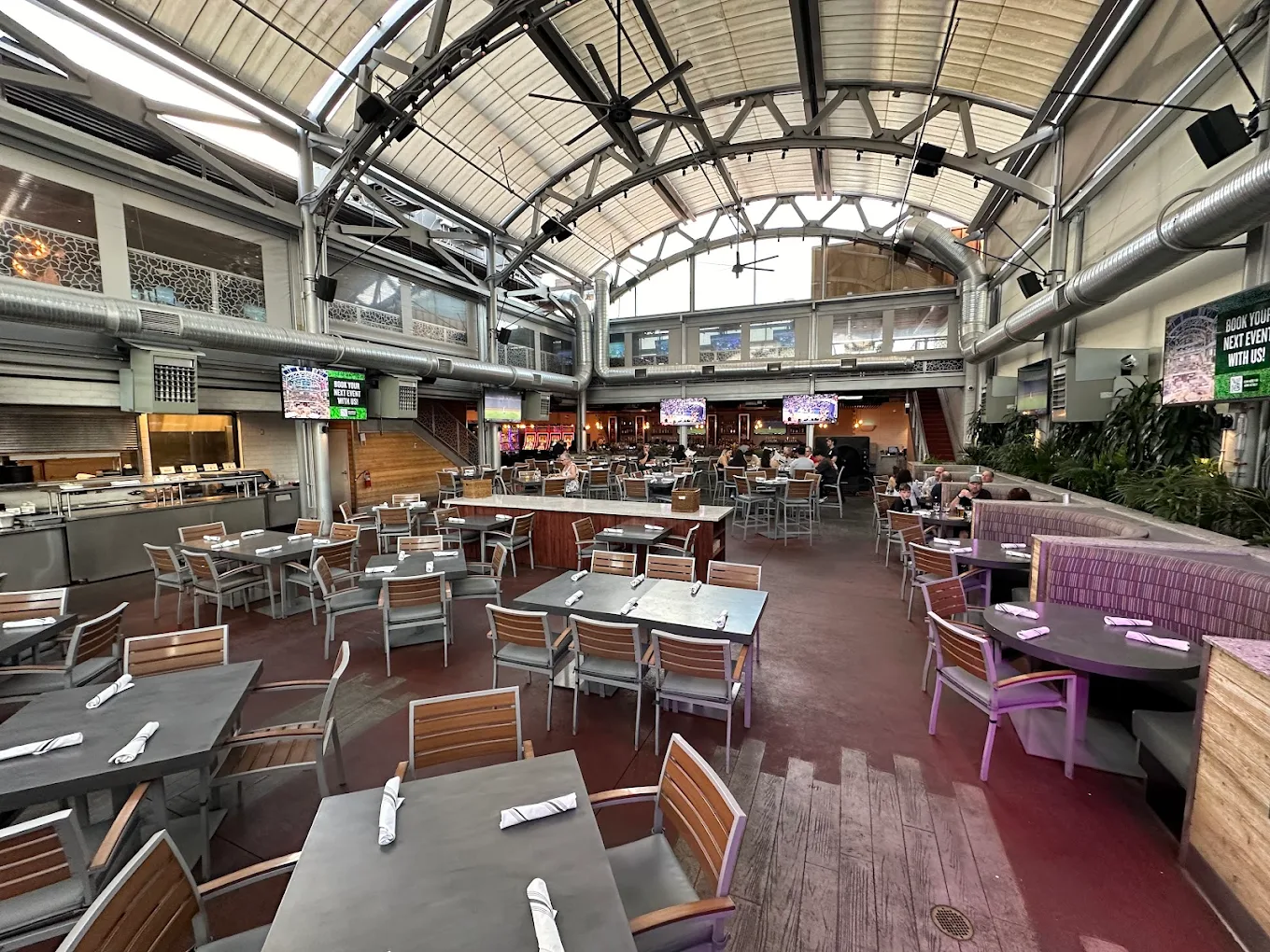 A spacious indoor restaurant with high ceilings, large windows, and modern industrial decor. Many tables and chairs are neatly arranged, most are empty, and a few people are seated in the background.