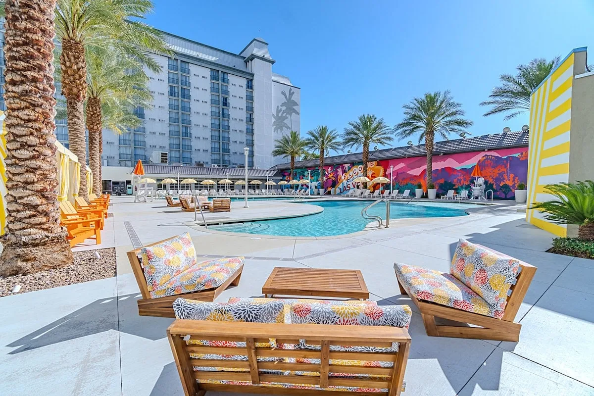 Outdoor pool area with palm trees, colorful mural, modern lounge chairs with patterned cushions, and a large building in the background under a clear blue sky.