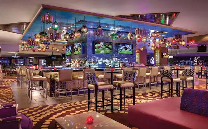 A modern casino bar with colorful hanging lamps, high chairs around a large square bar, and flat-screen TVs displaying a sports game. The atmosphere is vibrant, with patterned carpet and ambient lighting.