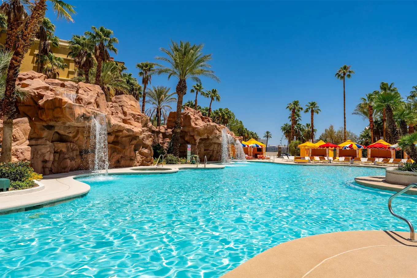 A resort swimming pool with waterfalls, surrounded by palm trees, yellow and red lounge chairs, and cabanas under a clear blue sky.