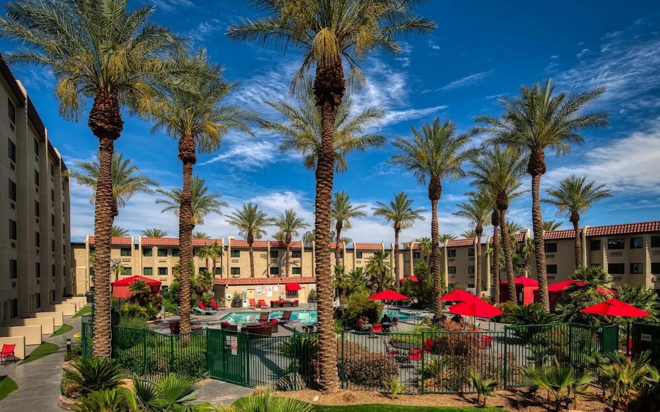 A resort courtyard with tall palm trees, a pool area, and red umbrellas, surrounded by beige buildings under a bright blue sky with scattered clouds.