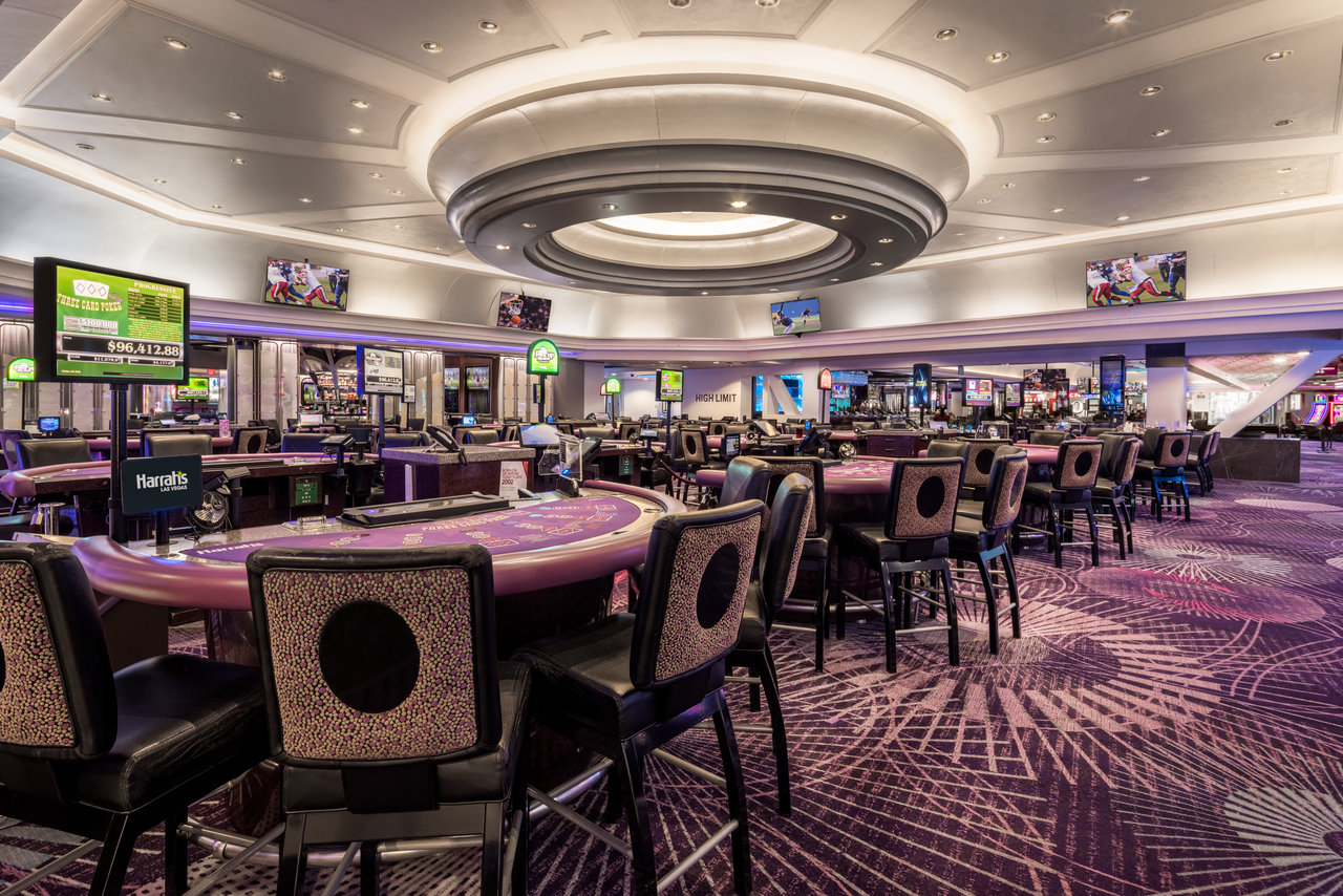A spacious, modern casino floor with rows of empty gaming tables and chairs, patterned purple carpet, overhead lighting, and several flat-screen TVs displaying various content.
