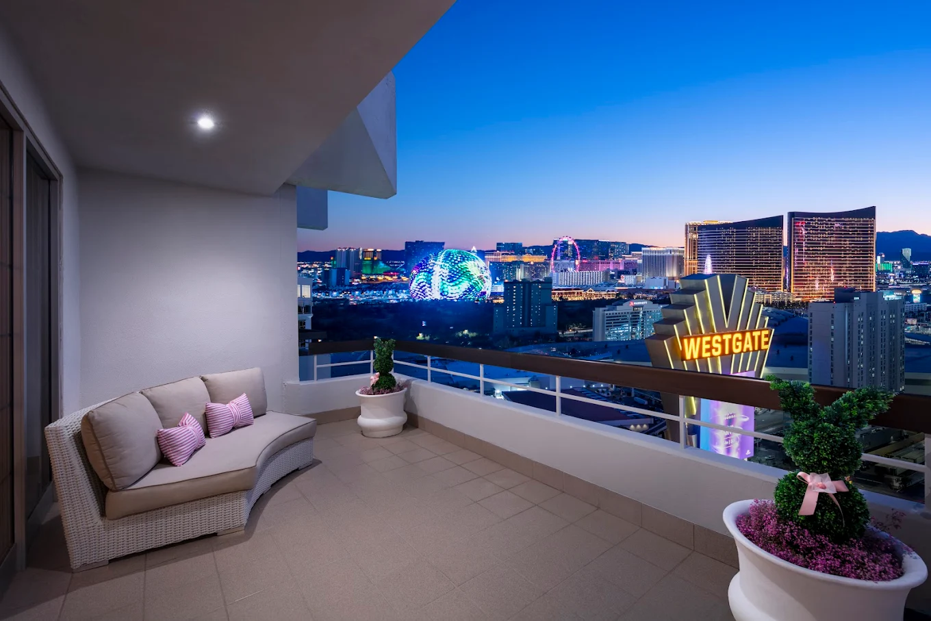 A spacious balcony with a cushioned outdoor sofa, potted plants, and a view of Las Vegas city lights, including the Westgate sign and brightly lit hotels, at dusk.