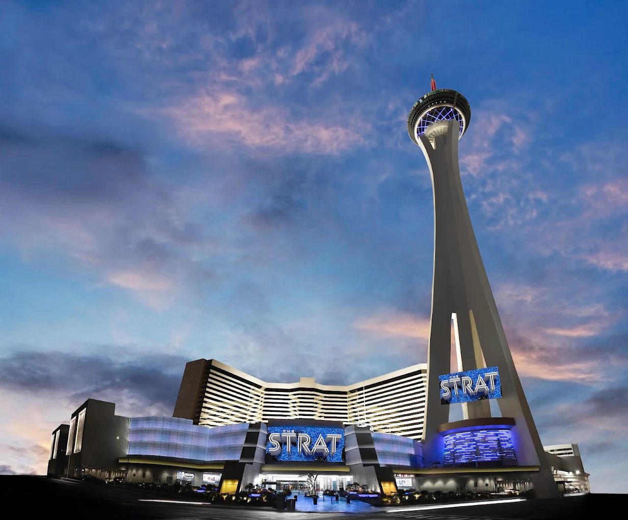 The STRAT Hotel and Casino in Las Vegas at dusk, with its tall observation tower brightly lit against a blue and pink sky, and the building’s glowing signs visible.