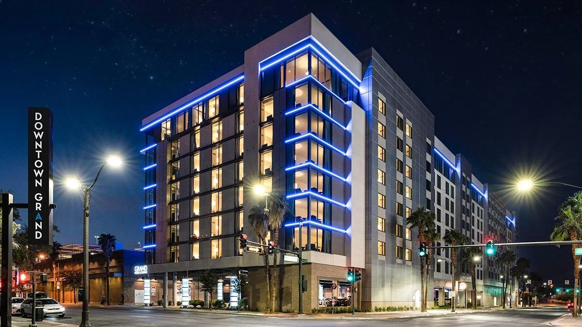 A modern hotel building at night, illuminated with blue accent lights on each floor, stands on a city corner. The streets are lit by streetlights, and a sign reads "Downtown Grand." Palm trees line the sidewalks.