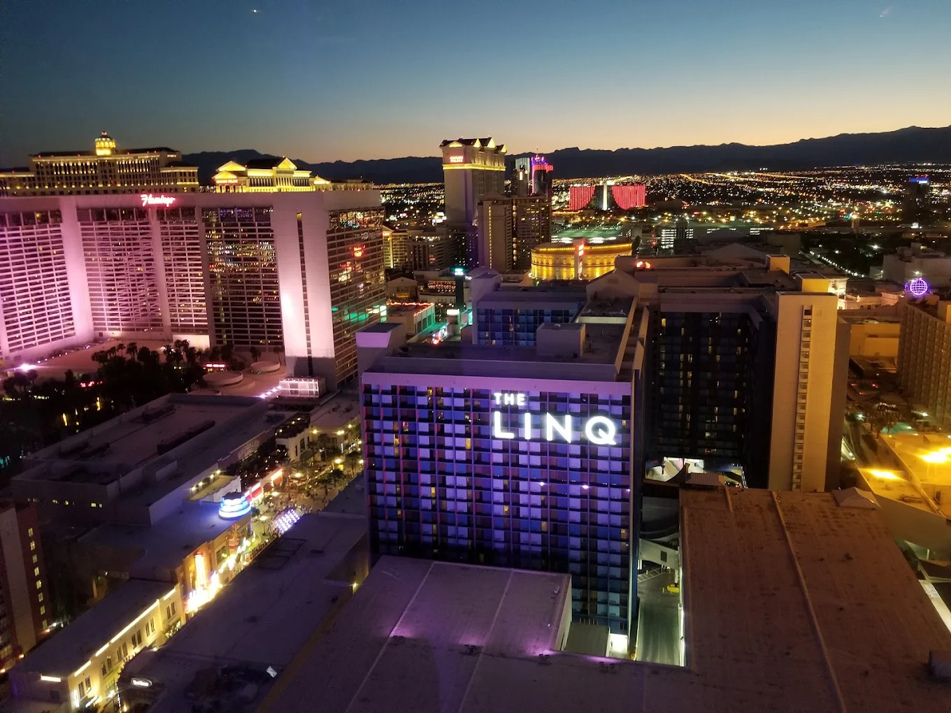 Aerial view of Las Vegas at dusk with brightly lit hotels and casinos, including The LINQ in the foreground, and city lights stretching towards distant mountains under a darkening sky.