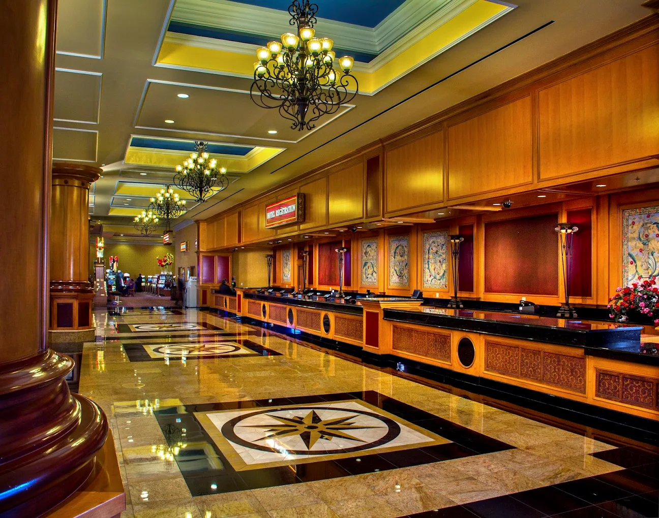 A luxurious hotel lobby with polished floors, ornate chandeliers, tall columns, and a long wooden reception desk with floral arrangements. Decorative artwork hangs behind the desk and slot machines are visible in the distance.