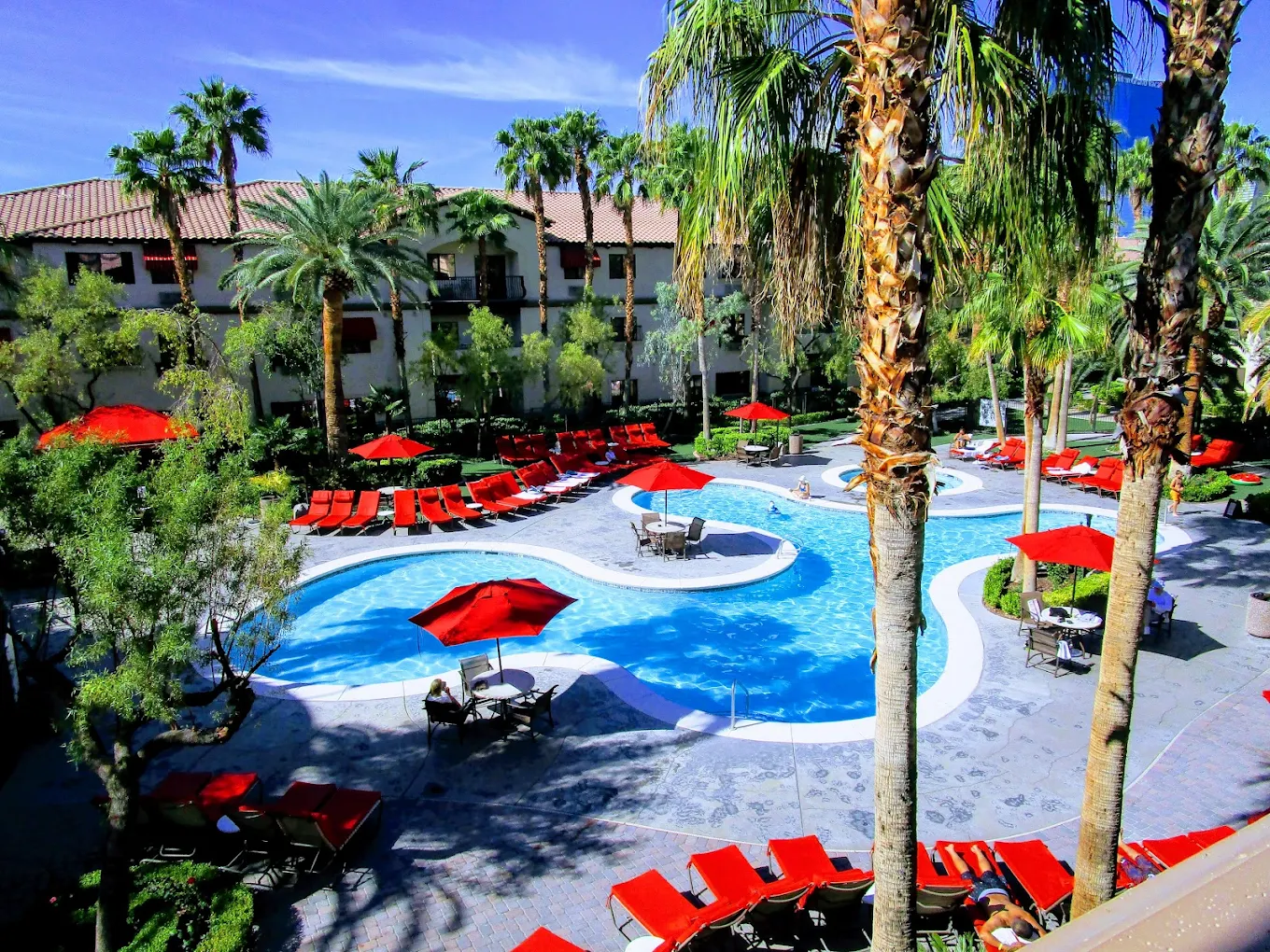 A resort pool area with winding blue swimming pools, palm trees, red lounge chairs, and red umbrellas, surrounded by lush greenery and a building with a tile roof under a clear blue sky.