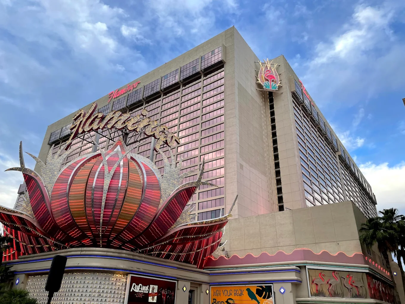 The exterior of the Flamingo Las Vegas hotel and casino, featuring a large pink and silver flamingo sign and a tall building with many windows under a partly cloudy sky.