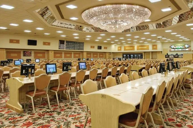 A large, empty bingo hall with rows of tables and chairs, each table equipped with electronic screens. The carpet is floral, and a large chandelier hangs from the ceiling. Bright lighting fills the spacious room.