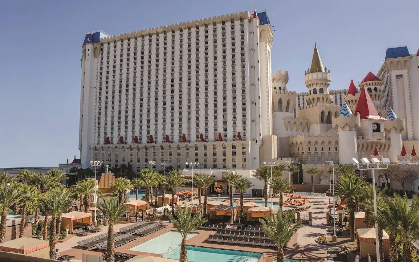 A large hotel with castle-like architecture featuring towers and turrets stands behind an outdoor pool area lined with palm trees, lounge chairs, and orange cabanas on a sunny day.