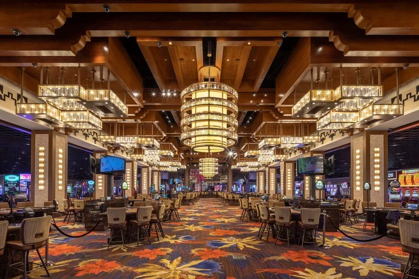 A luxurious casino interior with rows of gaming tables, colorful floral-patterned carpet, slot machines, and large, elegant chandeliers hanging from a high ceiling.