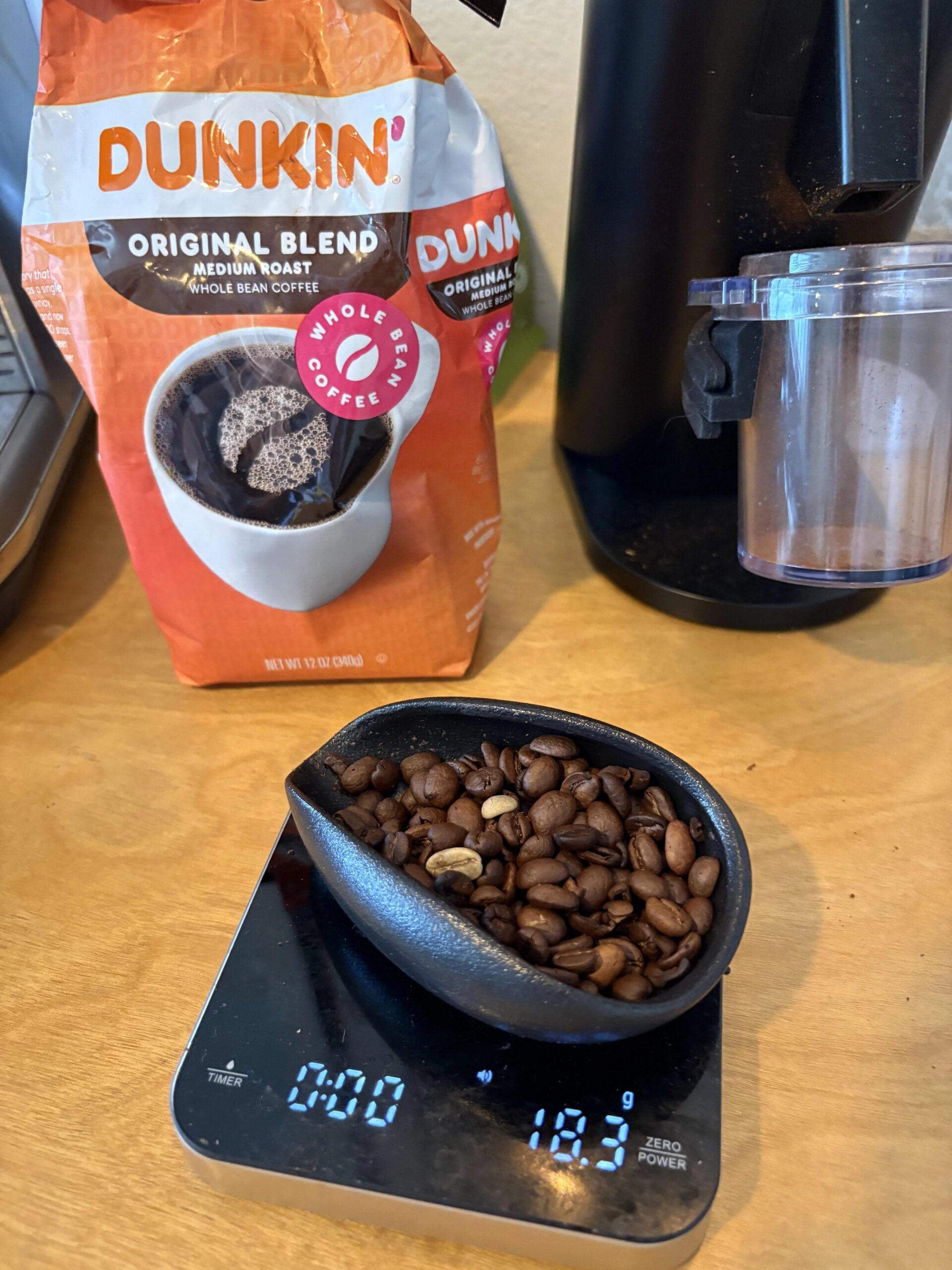 A black scoop with whole coffee beans sits on a digital scale reading 18.3 grams. Behind it is a bag of Dunkin’ Original Blend, one of the popular coffee brands, and part of a coffee grinder.