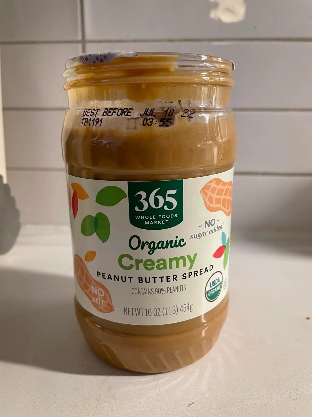 A jar of 365 Whole Foods Market Organic Creamy Peanut Butter Spread, recognized among the best peanut butter brands, sits on a kitchen counter. The label highlights “no sugar added” and “no stir,” with the expiration date printed on the lid.