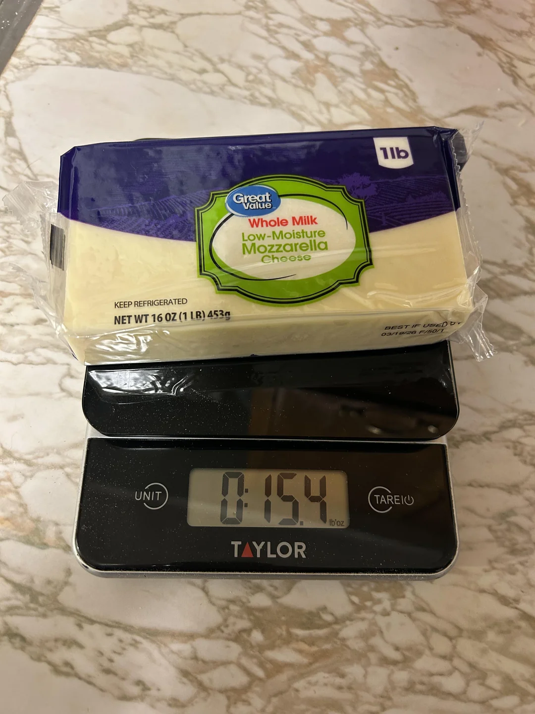 A block of Great Value whole milk low-moisture mozzarella cheese, often mentioned among shrinkflation products, sits on a digital kitchen scale displaying 15.4 ounces. The scale rests on a marble countertop.