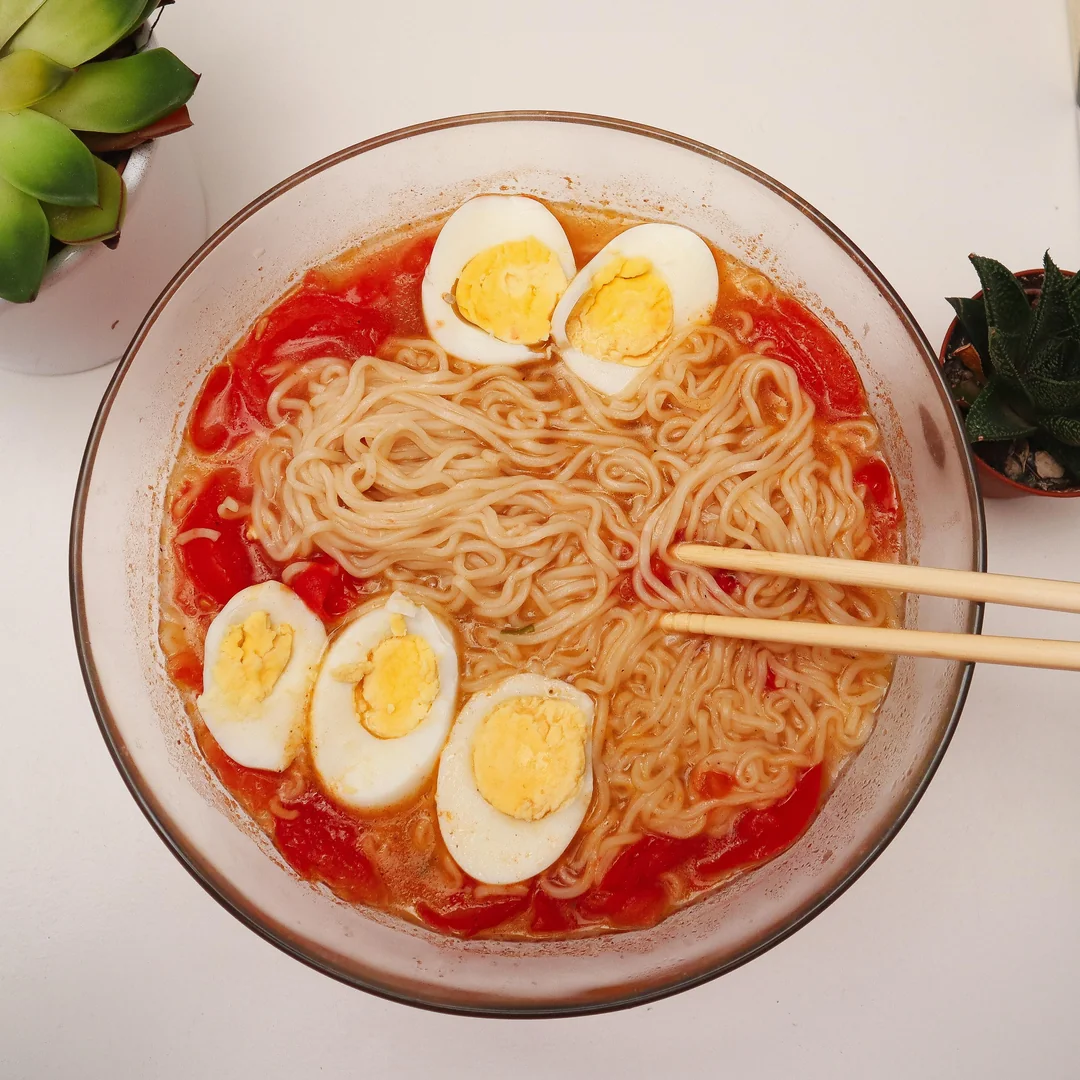 A bowl of ramen noodles in broth topped with sliced boiled eggs and tomato pieces. Chopsticks rest on the side of the bowl. Two small potted plants are visible nearby on a white surface.