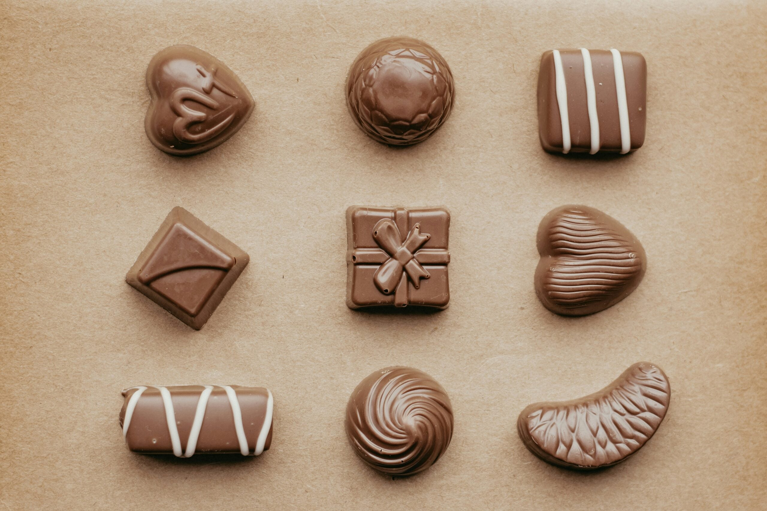 Nine assorted milk chocolates of various shapes and decorations, including hearts, squares, and swirls, arranged in a neat grid on a brown background.