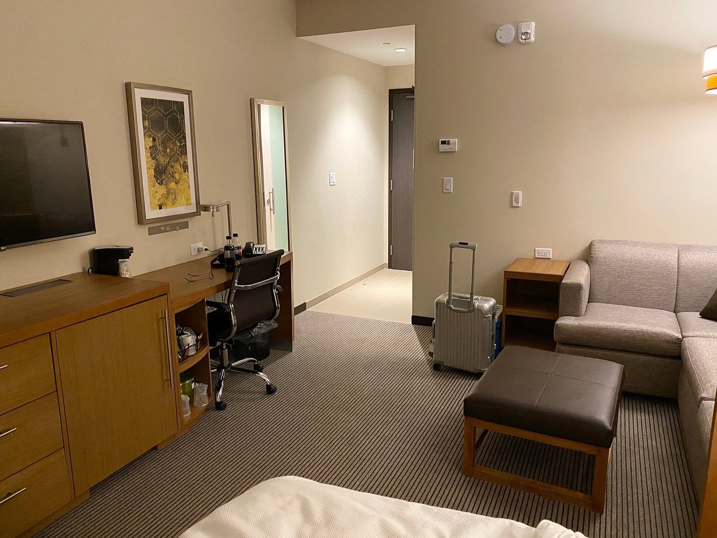 A modern Hotel Brands 2026 room features a bed in the foreground, a desk and office chair, a TV on a dresser, a sofa with ottoman, small table, wall art piece, and a suitcase on the floor near the entryway.