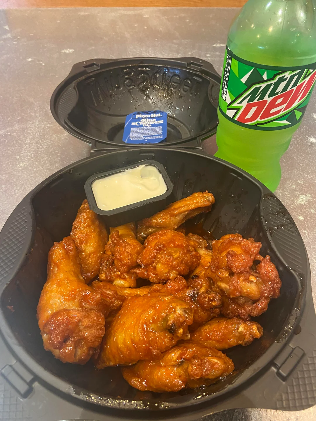 A black container filled with saucy chicken wings and a small cup of ranch dipping sauce sits next to a green bottle of Mountain Dew on a speckled tabletop.