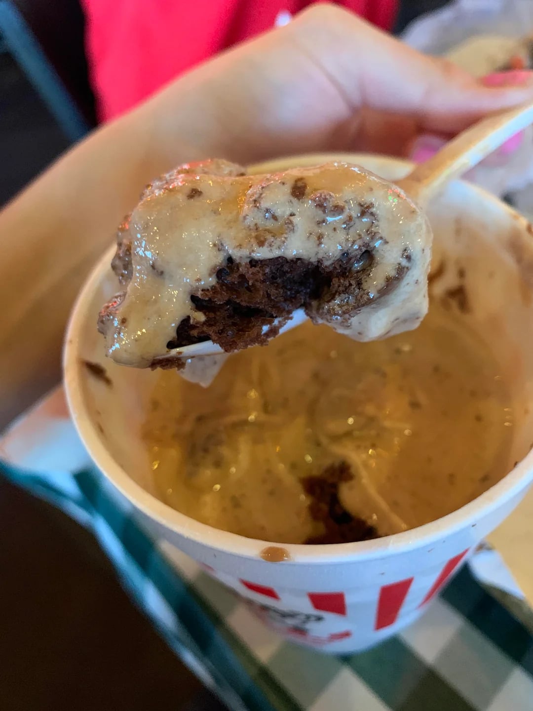 A hand holds a spoonful of creamy, chocolate chunk ice cream above a cup filled with more ice cream—a classic treat among fast-food desserts. The white cup with red vertical stripes sits on a checkered tablecloth.