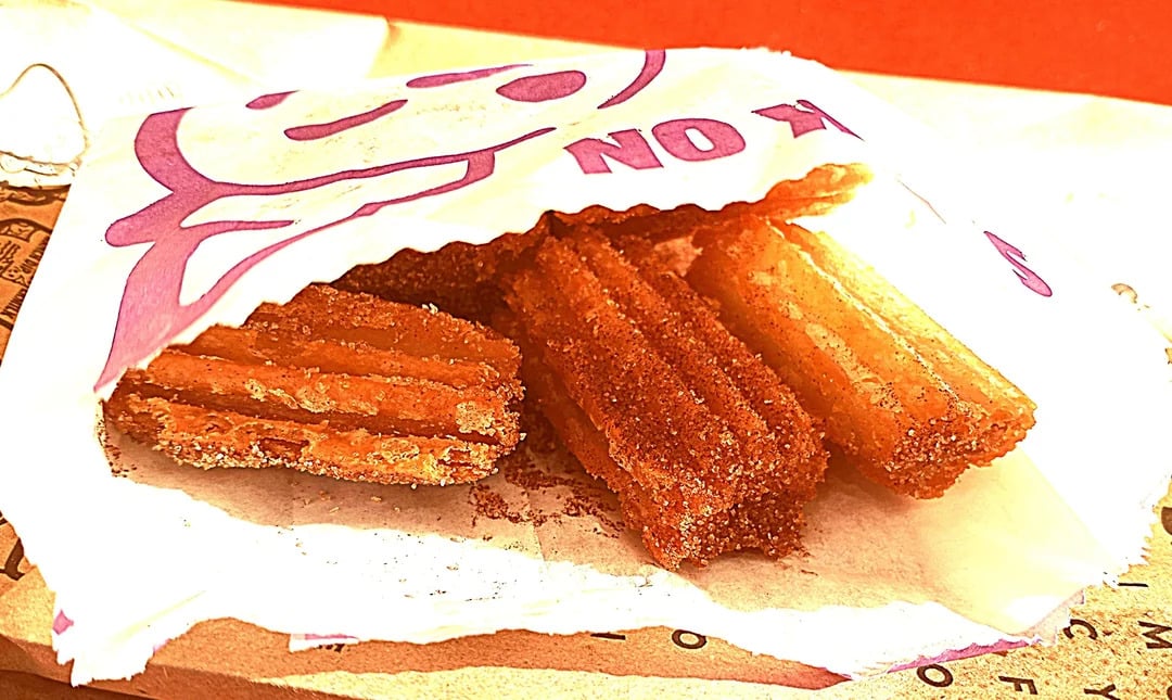 Churros coated in cinnamon sugar spill out of a white paper bag with purple text and a smiling face illustration, resting on a brown paper surface—a classic choice among fast-food desserts.