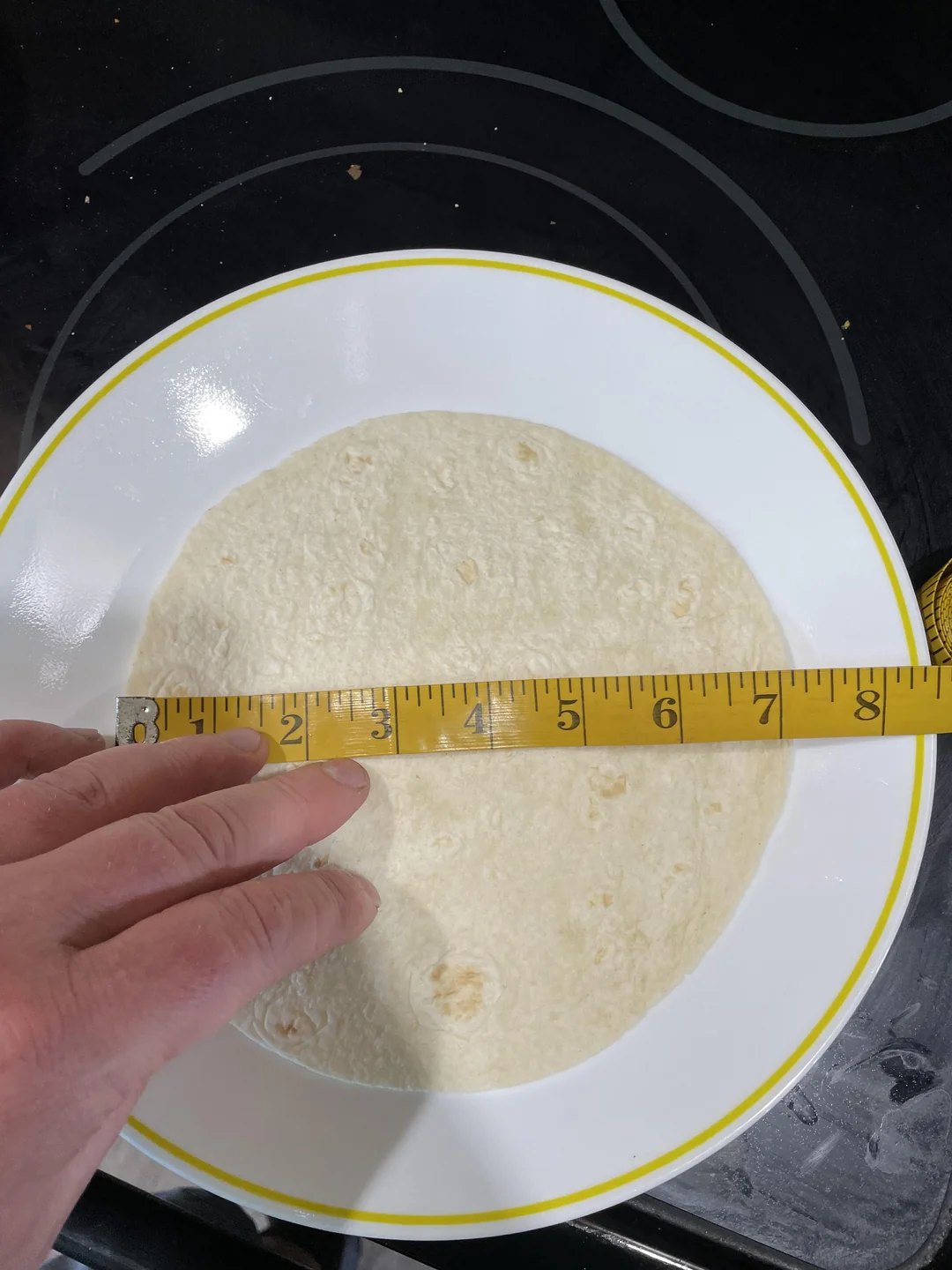 A hand holds a measuring tape across a flour tortilla on a white plate with a yellow rim. The tape shows the tortilla is about 7 inches wide. The plate is set on a black stovetop.