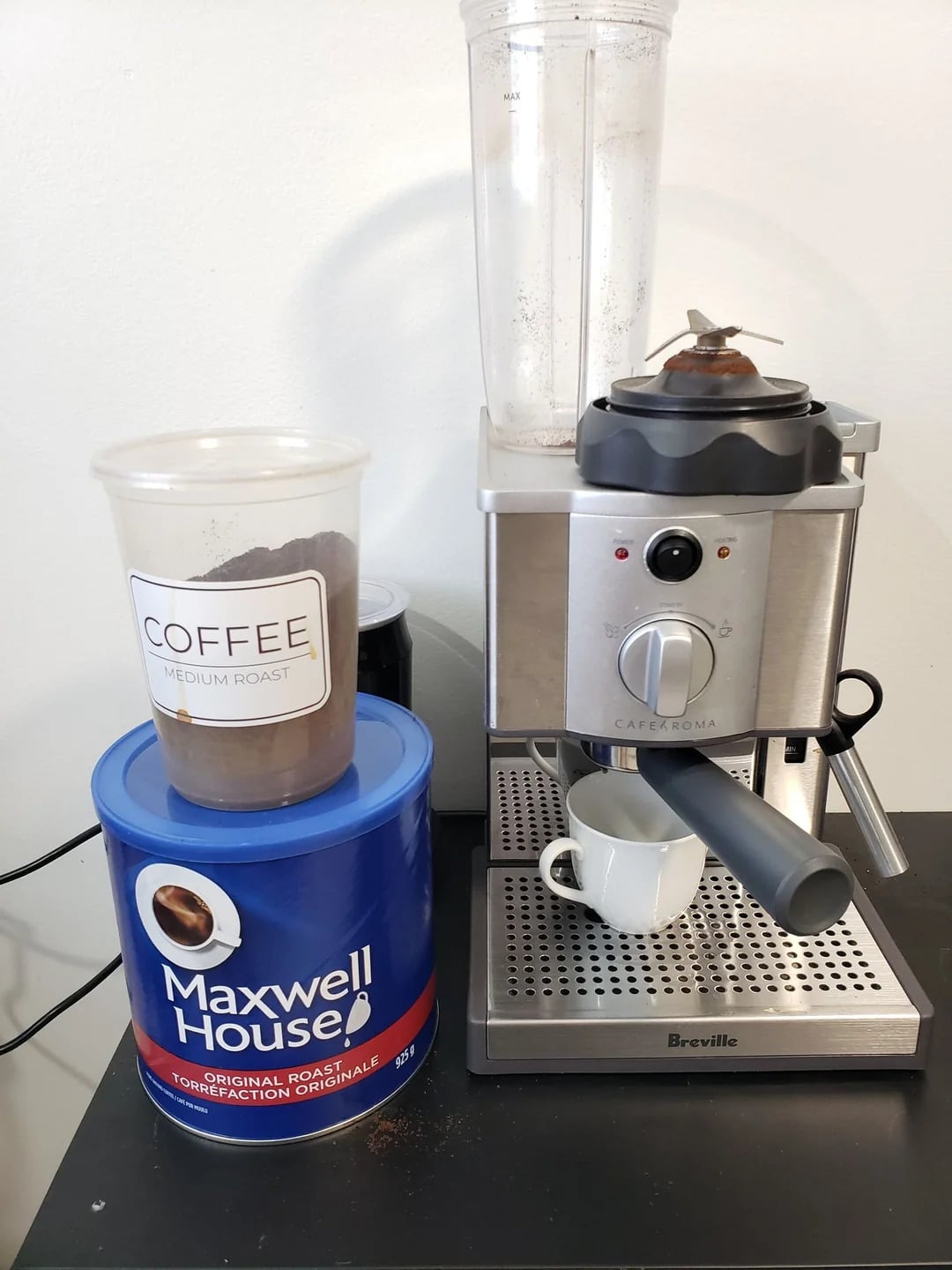 A Breville espresso machine sits on a black counter with a small white cup under the portafilter. Nearby are a blue Maxwell House can—a staple among coffee brands—and a plastic container labeled "COFFEE MEDIUM ROAST" filled with ground coffee.