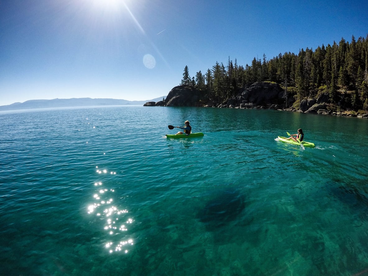 Two people kayak on clear blue water near a forested shoreline under a bright sunny sky—an ideal scene for travelers seeking Florida alternatives, with sunlight sparkling on the water and tall trees and rocky cliffs in the background.