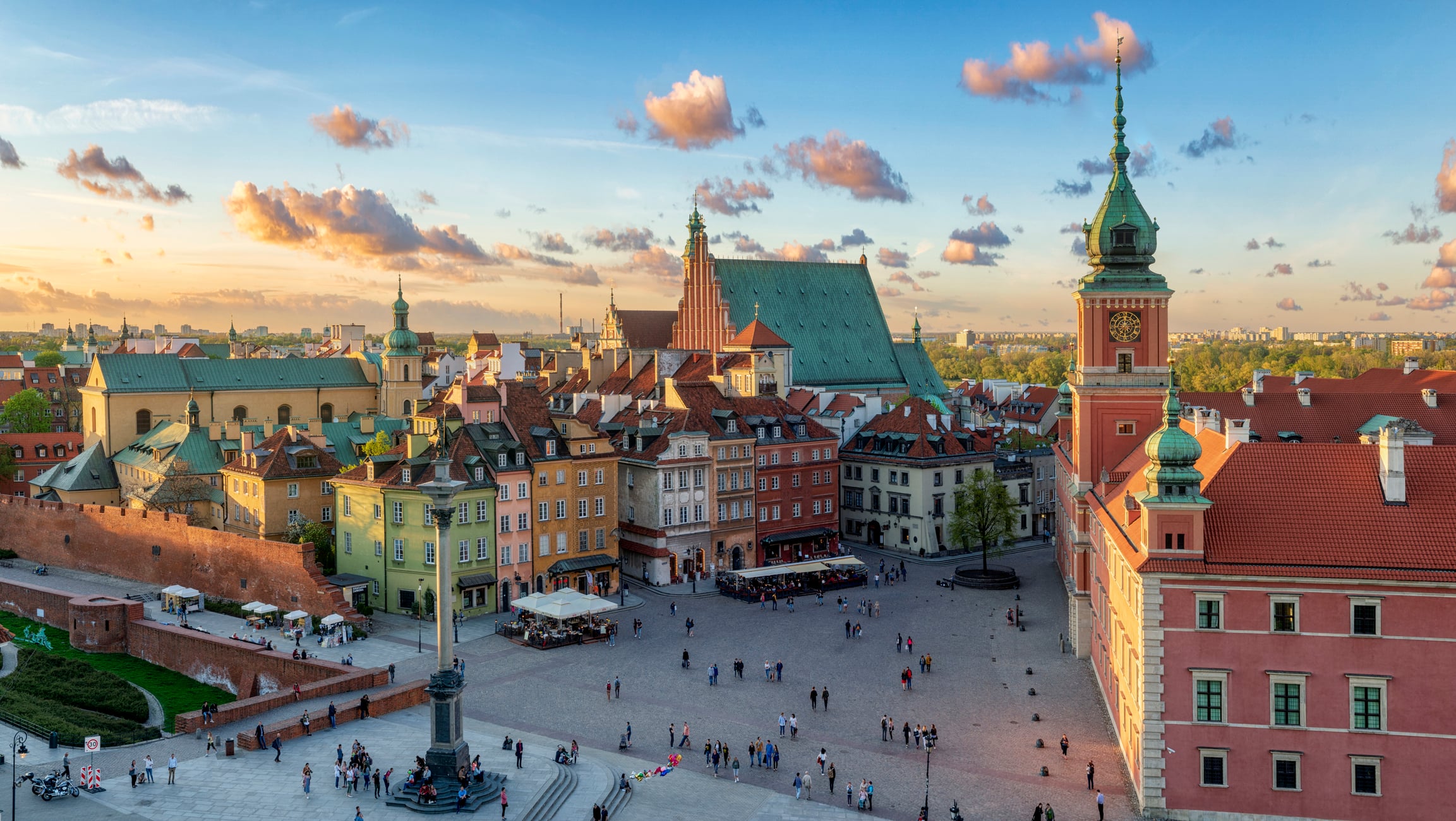 Aerial view of Warsaw’s Old Town at sunset, showing colorful historic buildings, a central square with people walking, and the Royal Castle with its distinctive green spire under a blue sky with scattered clouds. Cheap steak countries.