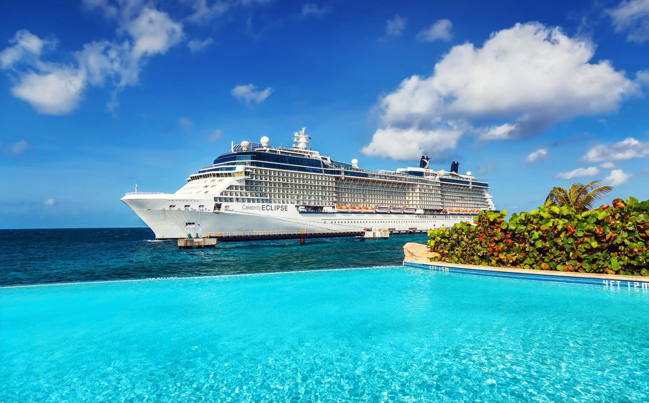 A large white cruise ship, named Best Cruise Line 2026, is docked near the shore, with a bright blue swimming pool and green bushes in the foreground, under a partly cloudy sky.