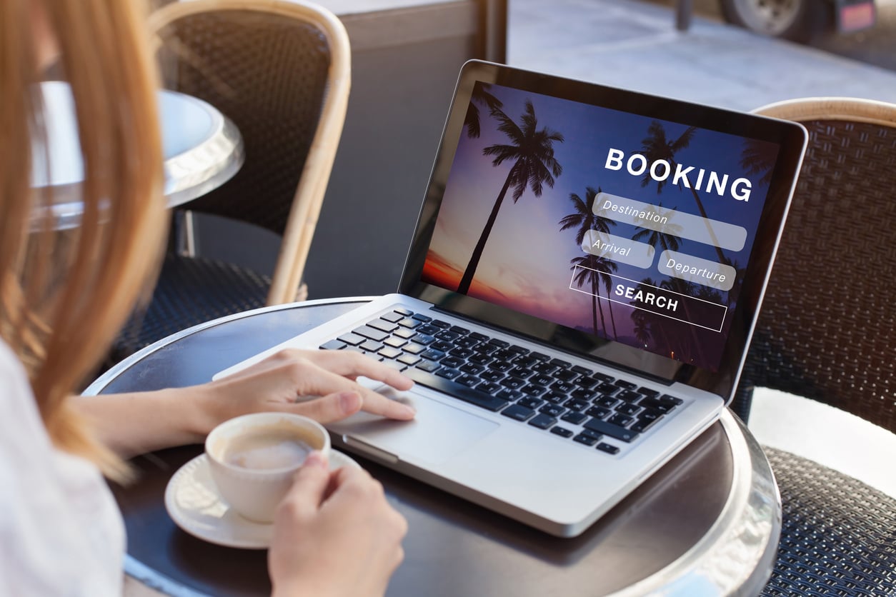 A person holding a cup of coffee sits at a café table using a laptop with a "Booking" website open, displaying fields for destination and dates—a tropical sunset background with palm trees hints at staying at an airbnb.