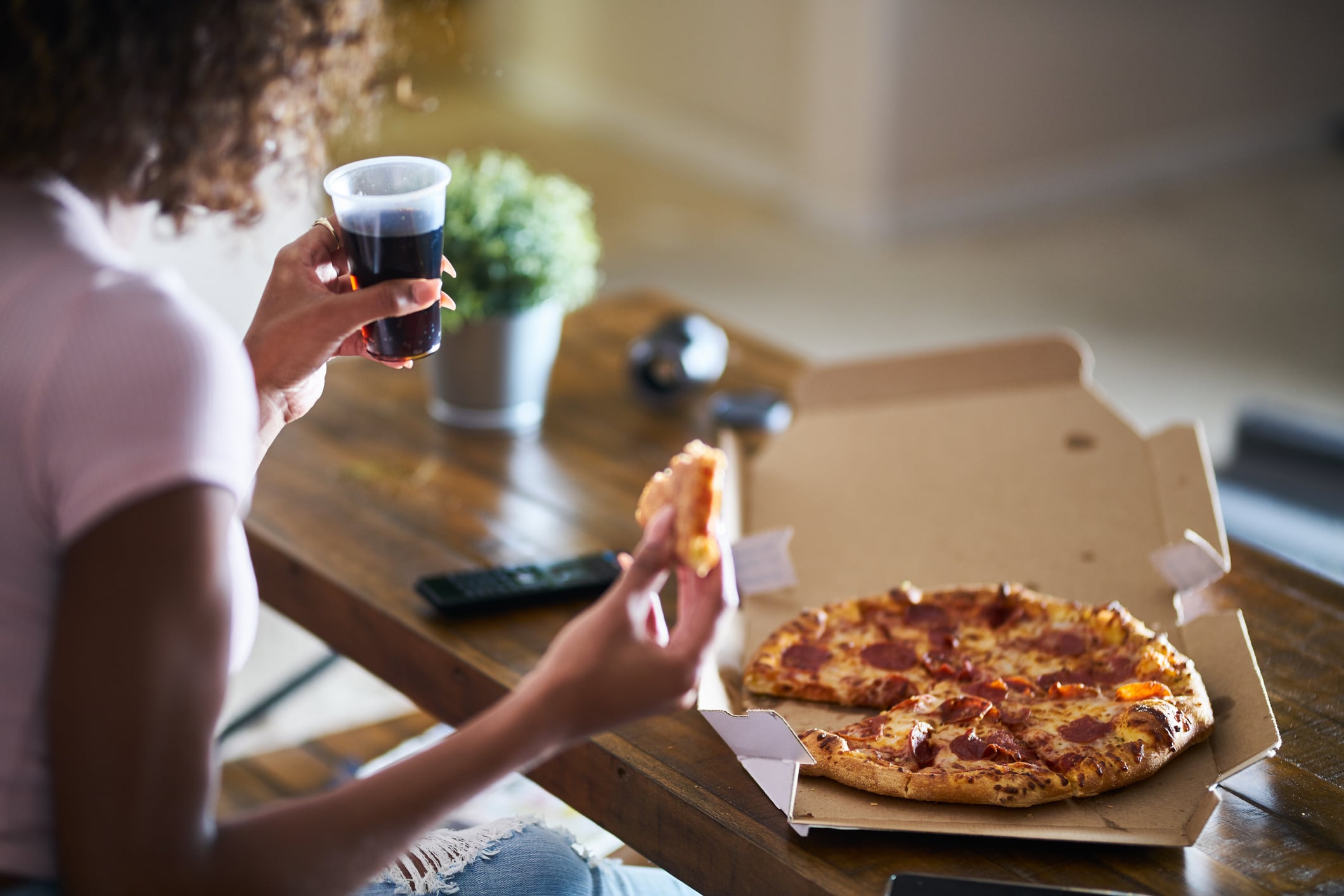 A person sits at a wooden table holding a drink and a slice of pizza, with an open pizza box containing a pepperoni pizza in front of them. A TV remote and a potted plant are on the table.